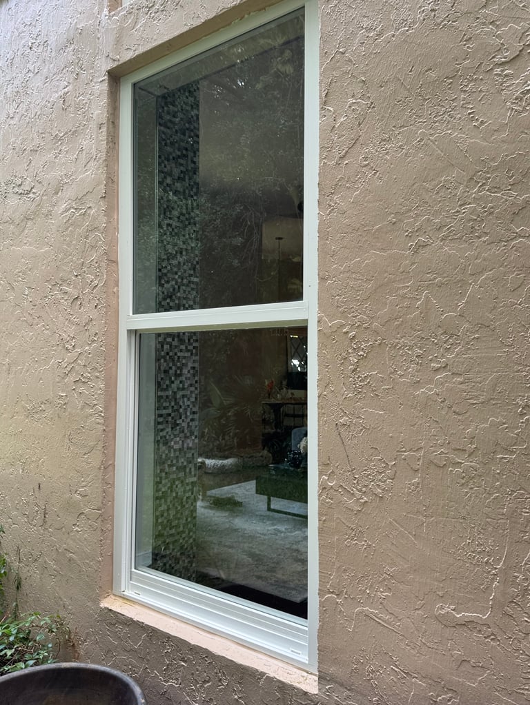 Double-pane window with white frame in textured beige stucco wall, reflecting interior patterned wall and room